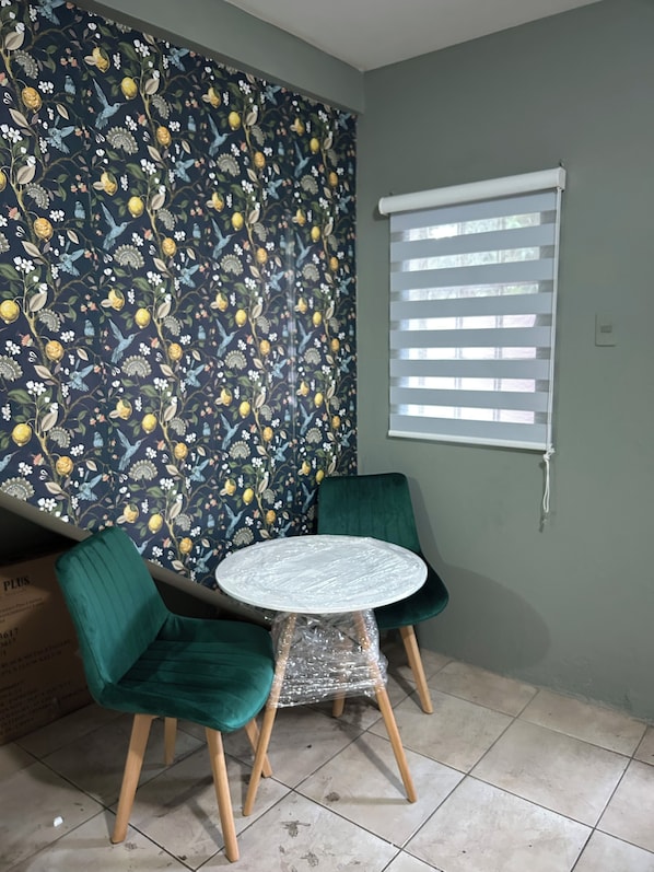 A room with a floral wallpaper, a round table with chairs, a window with blinds, and a green chair.