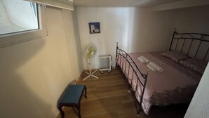 Une chambre avec un lit, un radiateur, un ventilateur sur pied, un petit banc et une fenêtre.