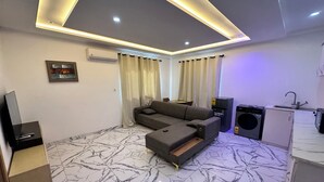 A modern living room with a sofa, a TV, a kitchen area with appliances, and a marble-patterned floor. - TN Residence  (Accra)