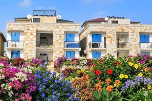 Un immeuble à plusieurs étages, doté de balcons et d’une façade en pierre, avec une variété de fleurs aux couleurs vives au premier plan.