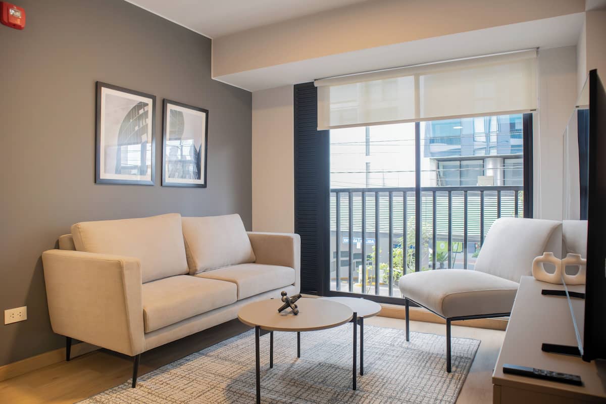 Uma sala de estar moderna com sofá, poltrona e mesa de centro, uma janela grande com persianas e quadros na parede.