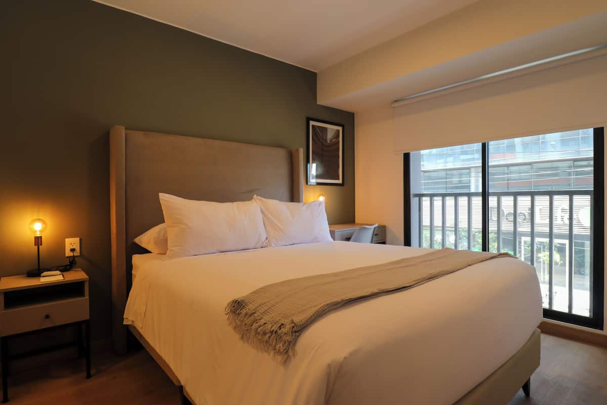 Quarto de hotel com uma cama grande, uma mesa de cabeceira com luminária, um quadro na parede e uma janela com vista para edifícios.