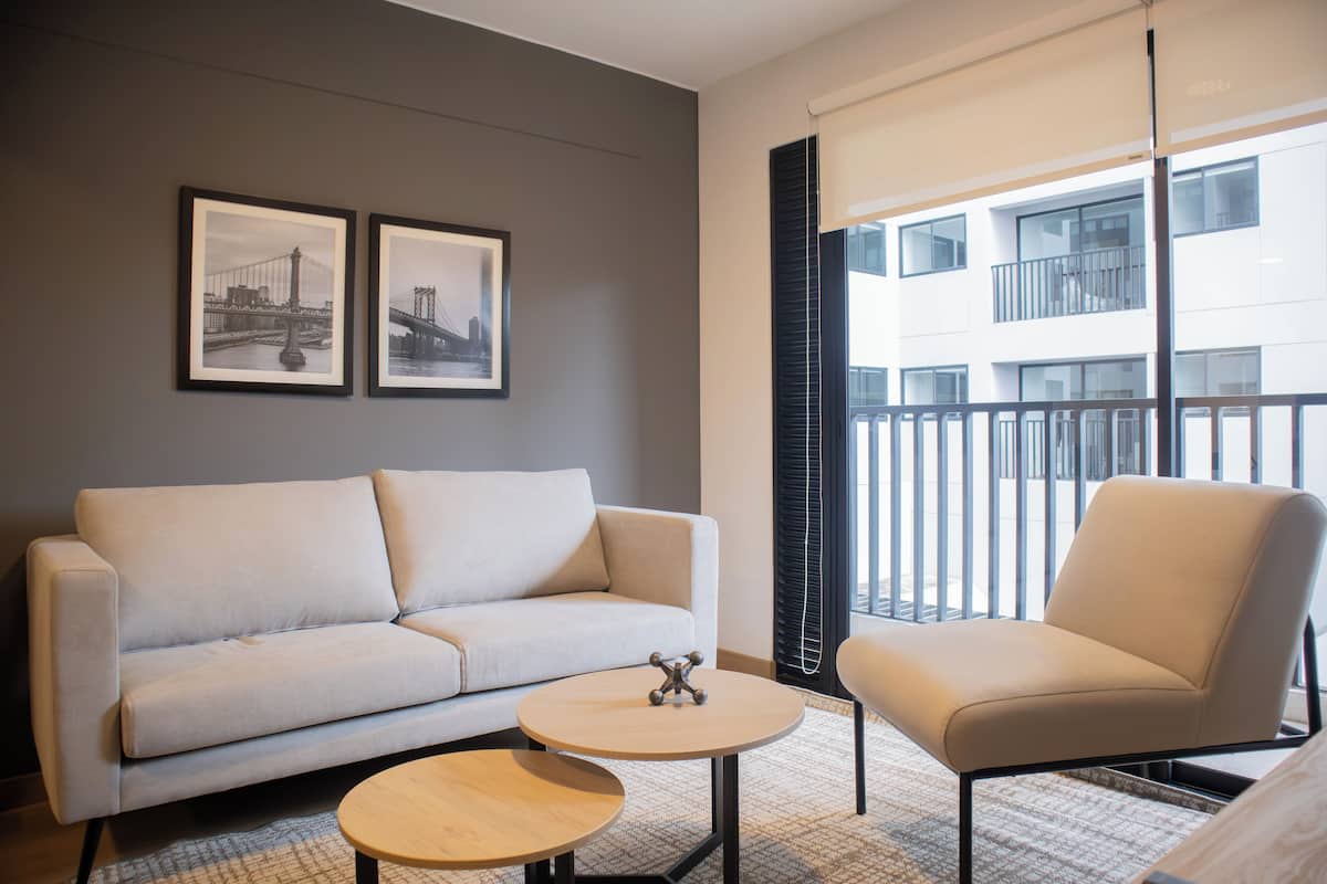 Sala de estar moderna com sofá, poltrona e mesa de centro. Há duas quadros nas paredes, uma janela com persianas e uma varanda com guarda-corpo.