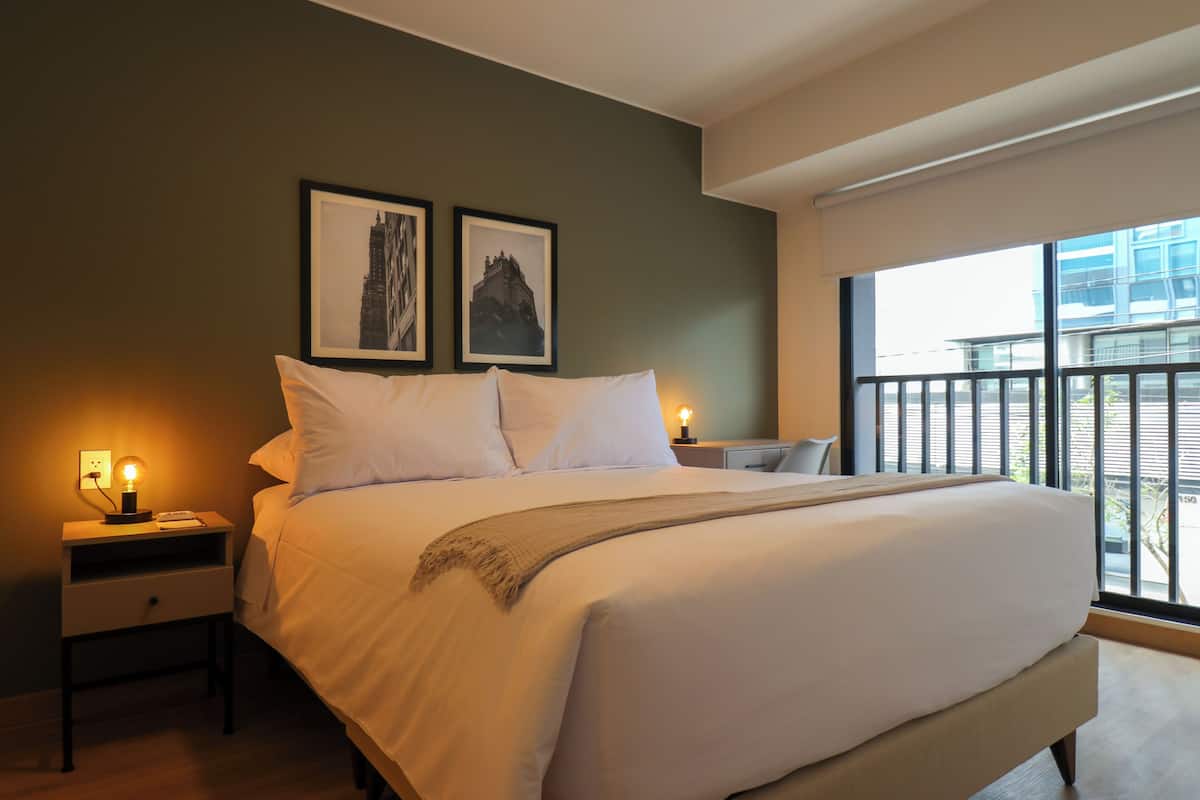Quarto de hotel moderno com uma cama grande, duas mesas de cabeceira e uma janela com vista para a cidade.