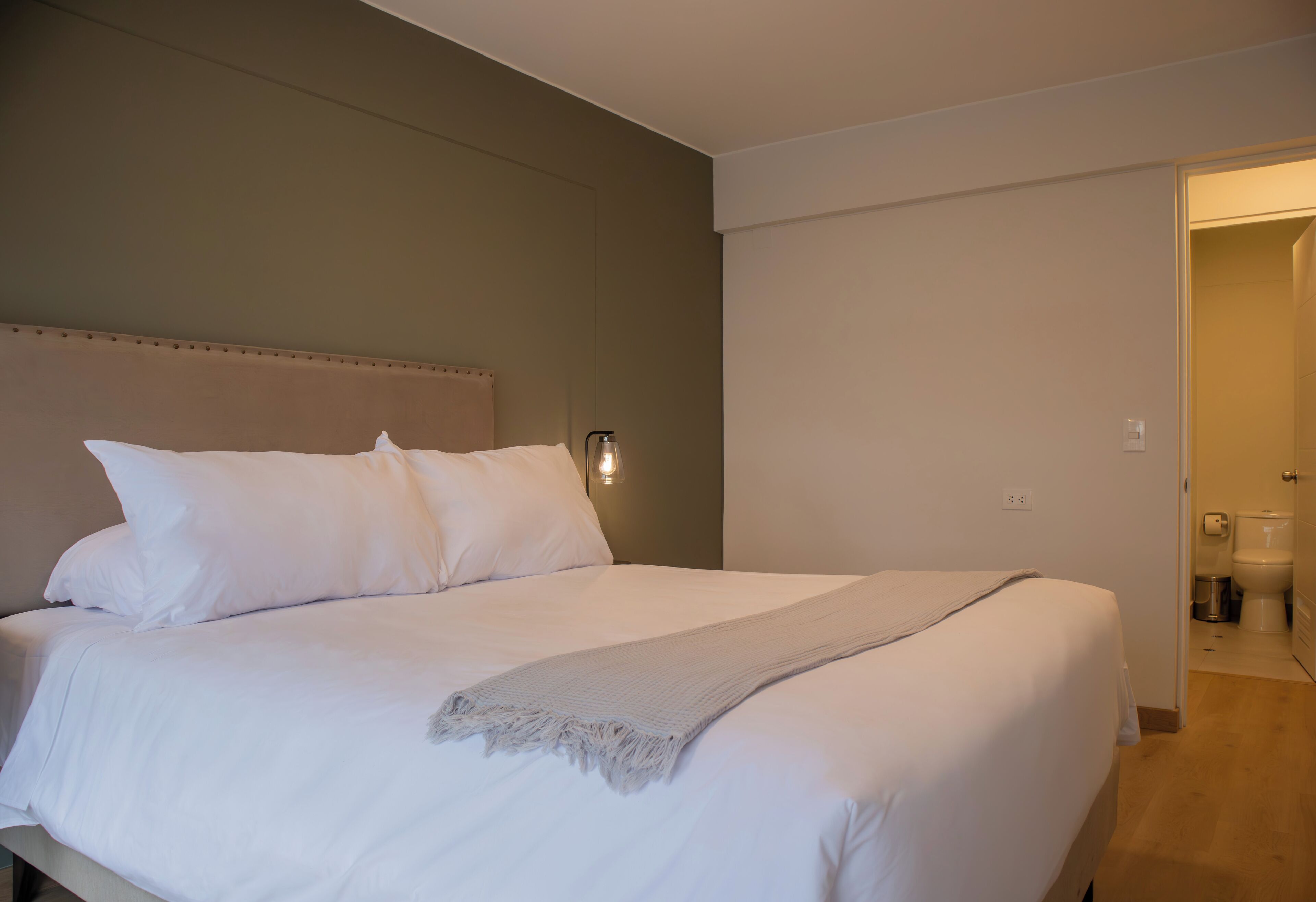 Un lit bien fait, avec du linge de lit blanc et un plaid gris, un luminaire fixé au mur, et une salle de bain visible, équipée de toilettes et d’une poubelle.