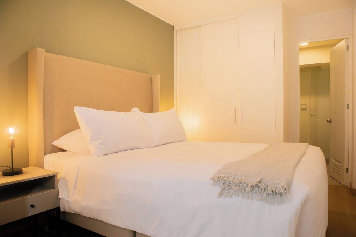 Cama bem arrumada com lençóis e almofadas brancos, mesa de cabeceira com abajur e guarda-roupa com portas deslizantes.