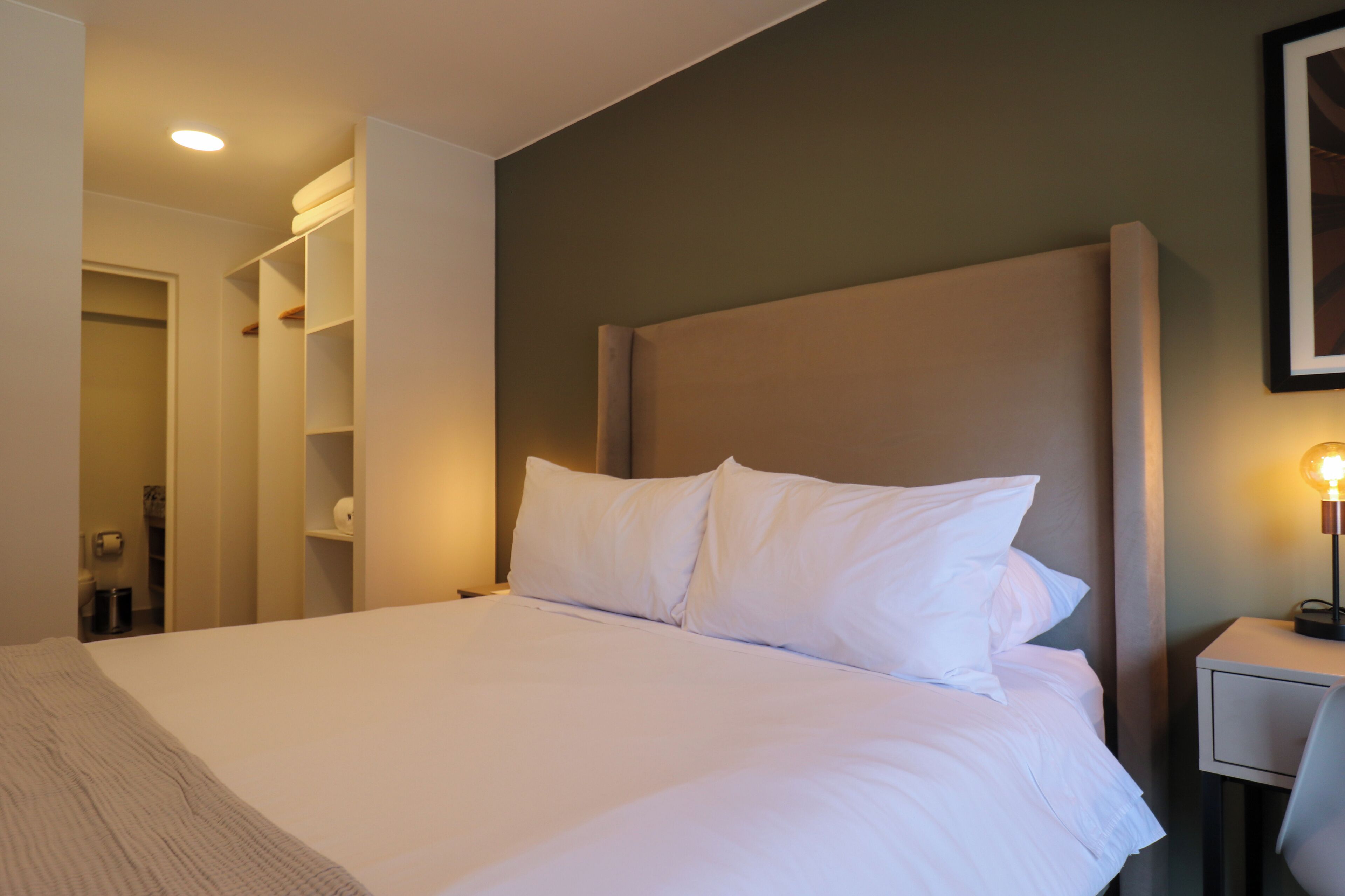 Quarto de hotel com uma cama grande, uma mesa de cabeceira com abajur e um guarda-roupa com prateleiras.