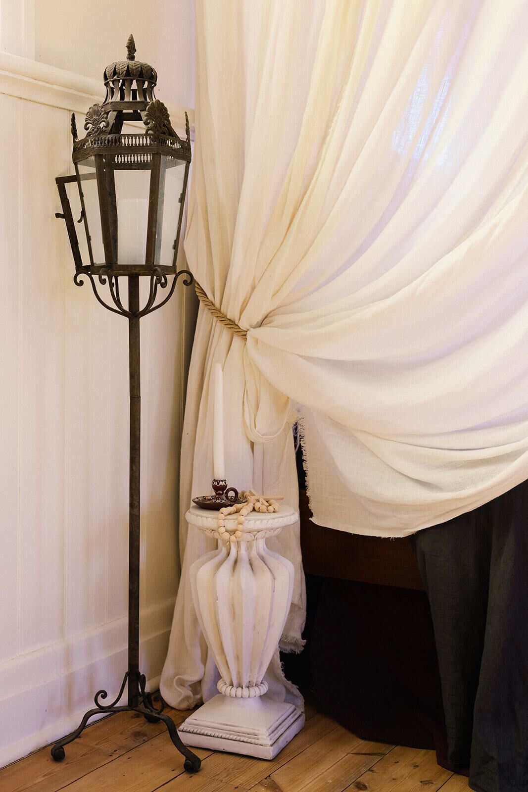 Een hoek met een witte gordijn, een wit voetstuktafeltje met een decoratief object en een zwarte metalen vloerlamp.