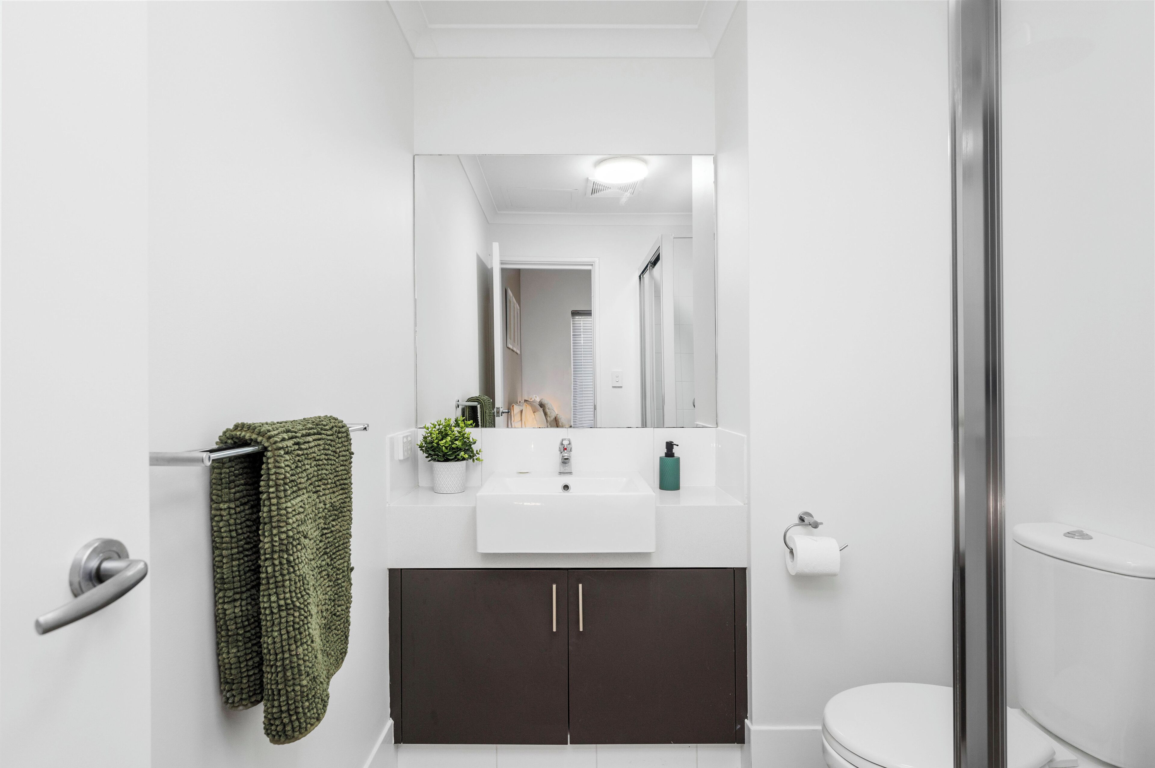 A modern bathroom with a white sink, dark cabinets, a green towel, a potted plant, and a toilet.