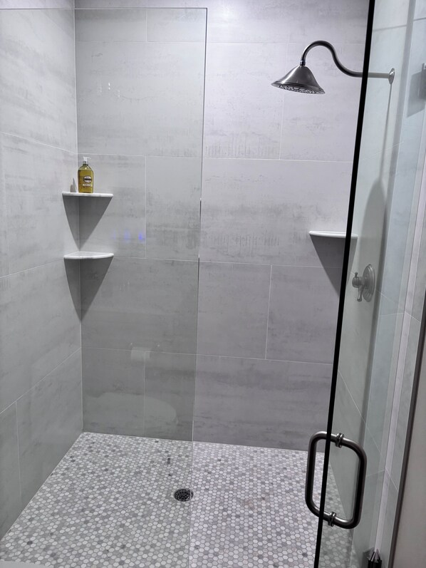 Un espace douche avec des carreaux gris, une douchette et un flacon de shampoing posé sur une étagère.