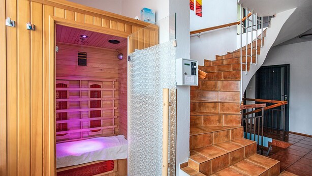 A sauna with a wooden door and a bench inside, a staircase with wooden steps and metal railings, and a white wall with a thermostat.