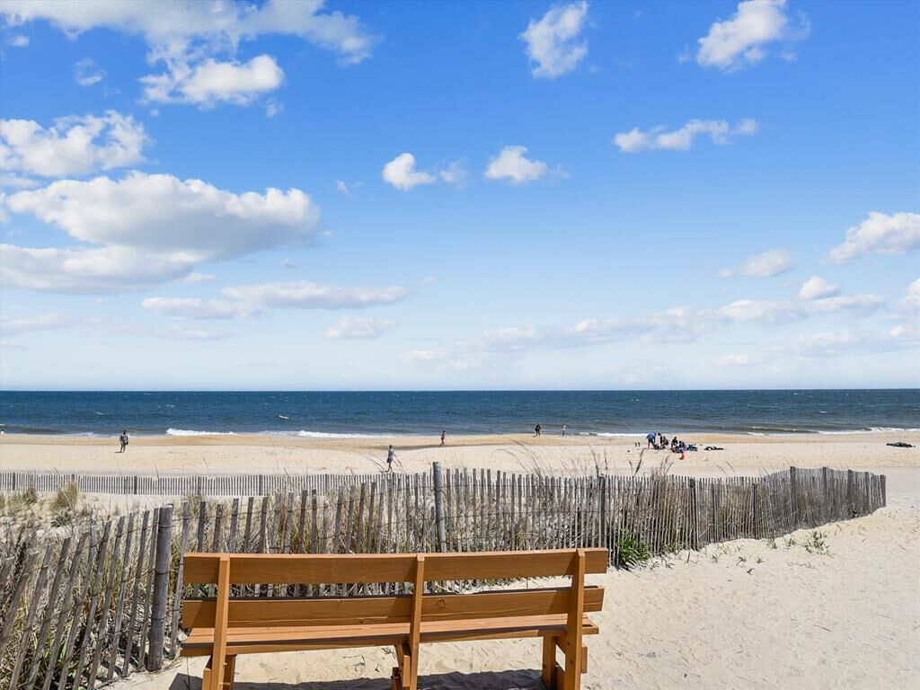 Un banc en bois sur une plage de sable, avec une clôture en bois en arrière-plan et un ciel bleu dégagé au-dessus.