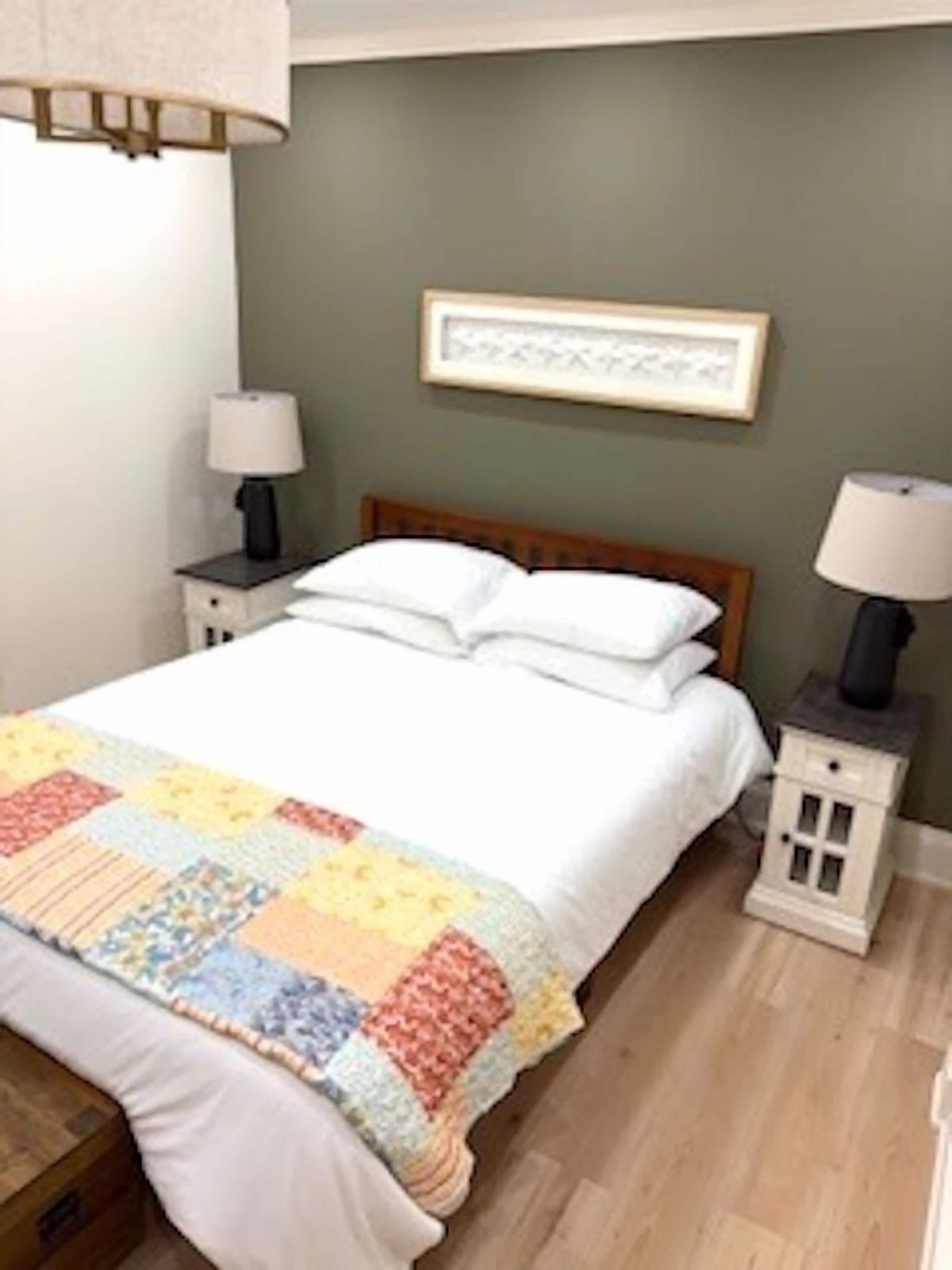 Una cama bien tendida con un edredón colorido, dos mesitas de noche con lámparas, un cuadro en la pared y una mesita auxiliar de madera.