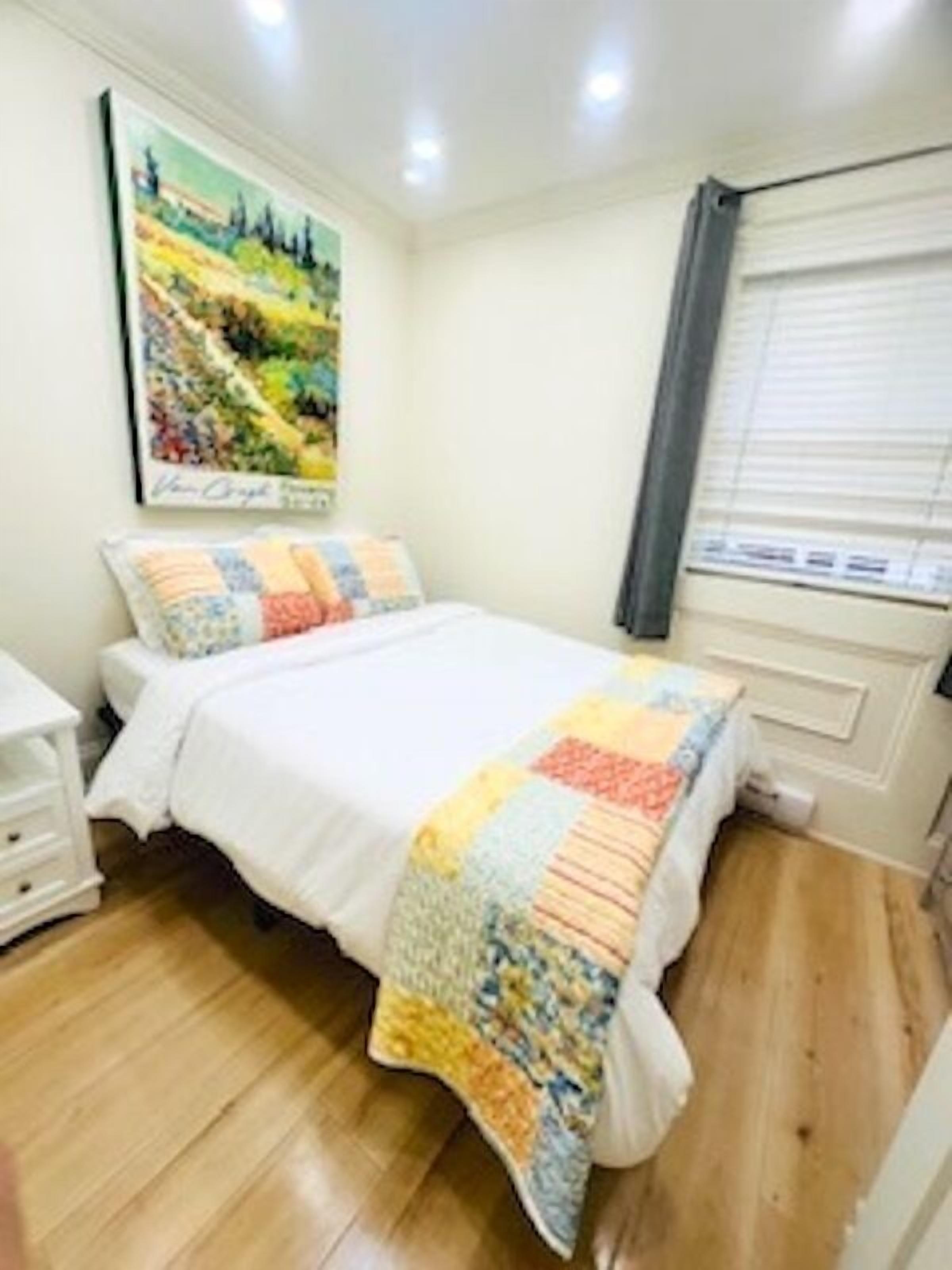A bedroom with a bed, a nightstand, a window with blinds, and a painting on the wall.