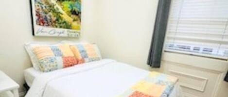 A bedroom with a bed, a nightstand, a window with blinds, and a painting on the wall.