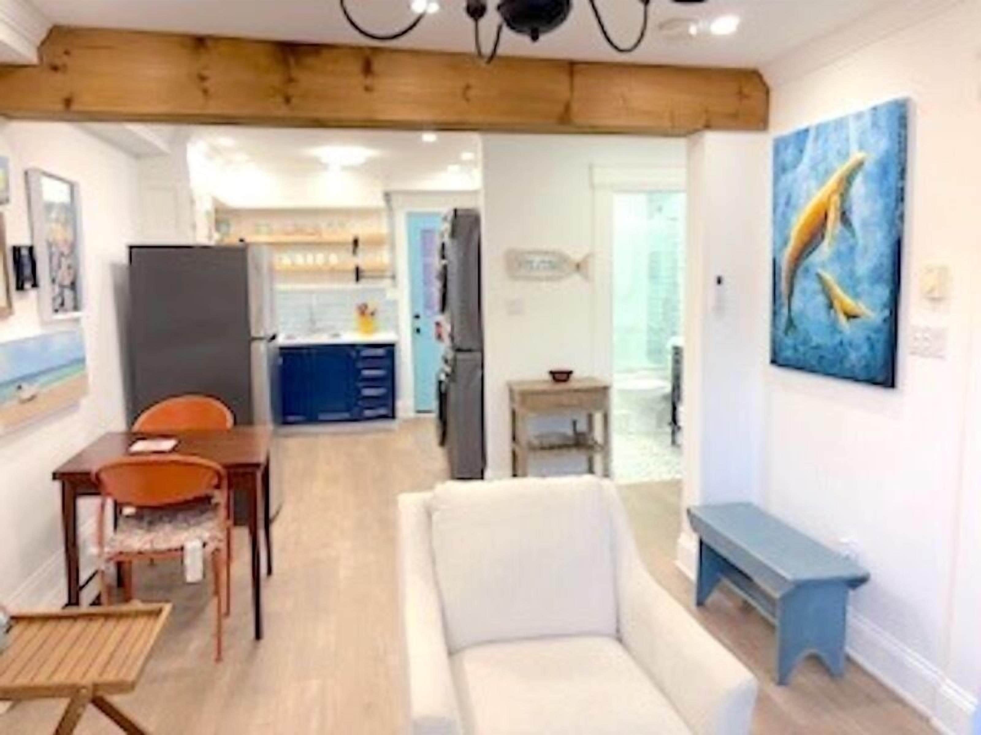 A living room with a white sofa, a wooden dining table with chairs, a small blue bench, and a painting of dolphins on the wall.