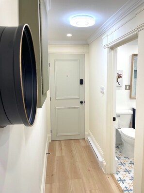 A hallway with a white door, a light fixture above, and a bathroom visible through an open door. - HarbourGate Condos & Studios (St. John's)