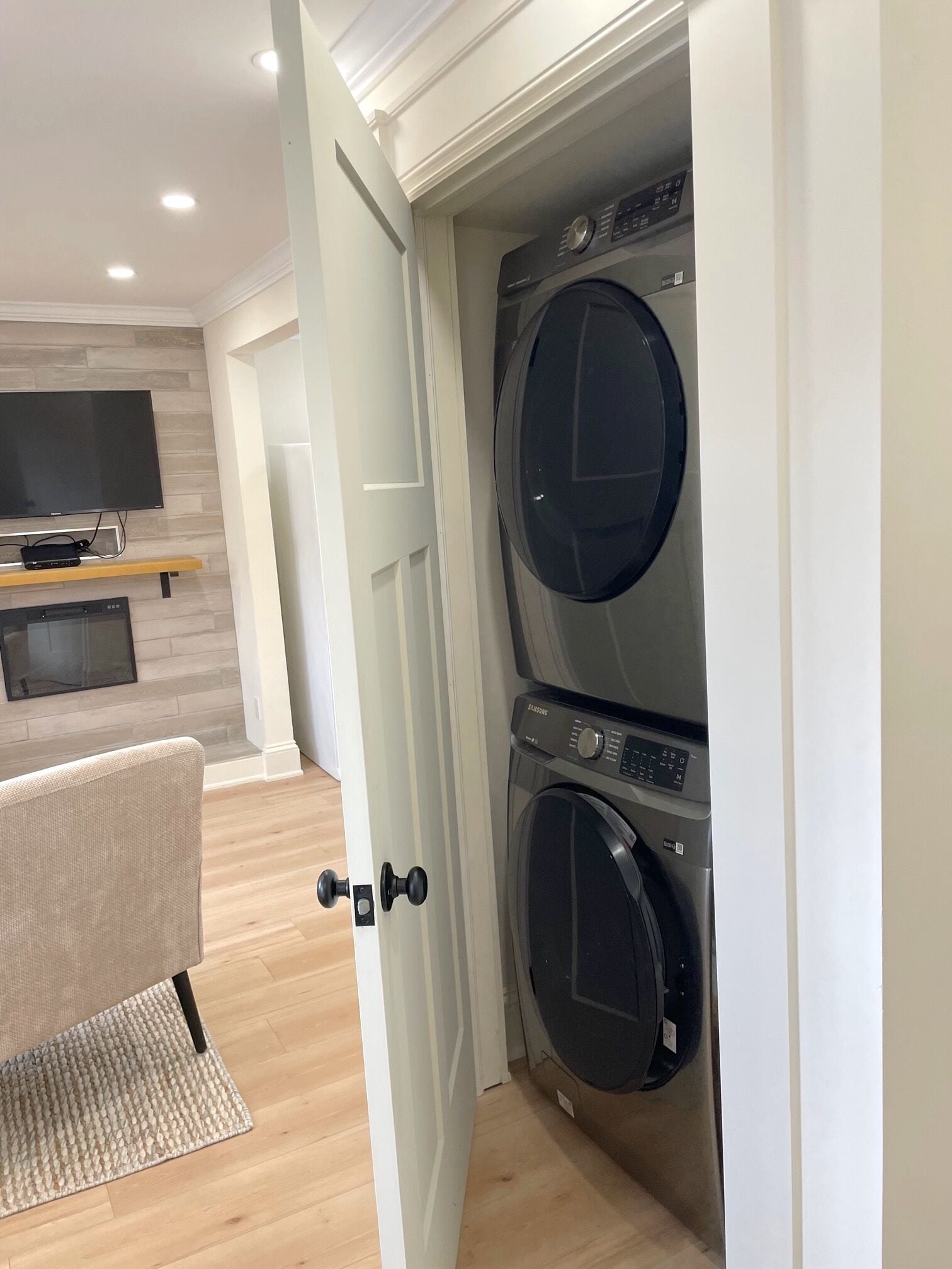 A laundry room with two stacked front-loading washing machines, a flat-screen TV on a wooden shelf, and a beige chair with a woven rug.