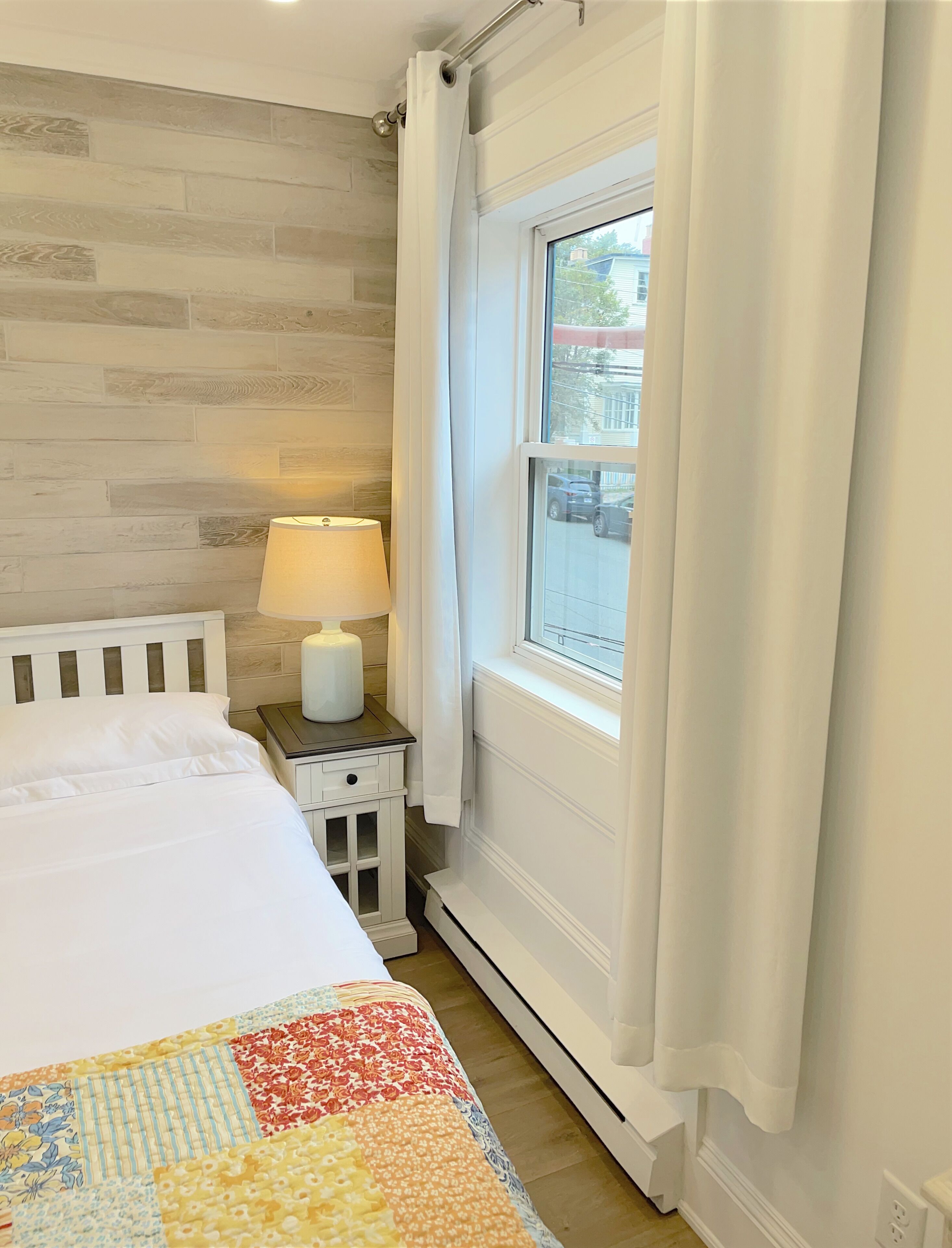 Une chambre avec un mur en bois, une fenêtre ornée de rideaux blancs, une lampe de chevet et une courtepointe sur le lit.