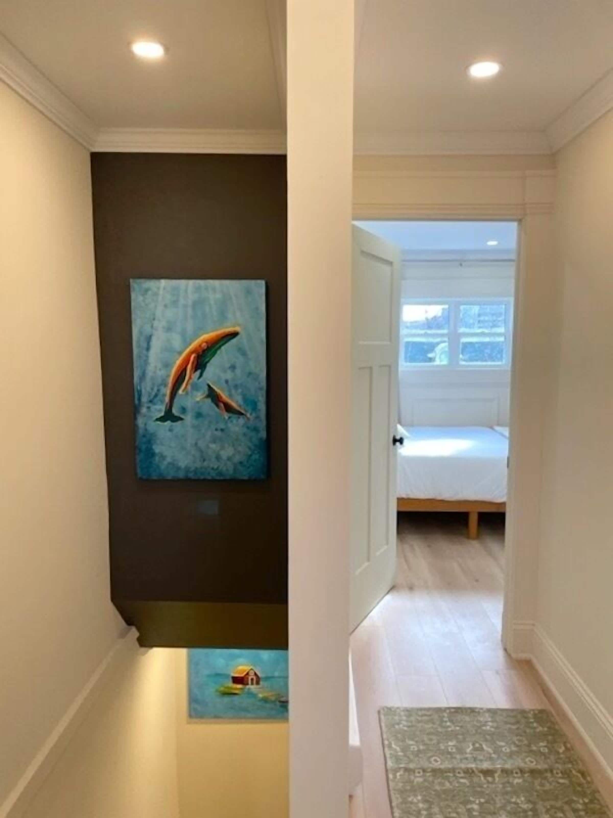 A hallway with a painting of dolphins, a small painting of a house on water, and a bed in the adjacent room.