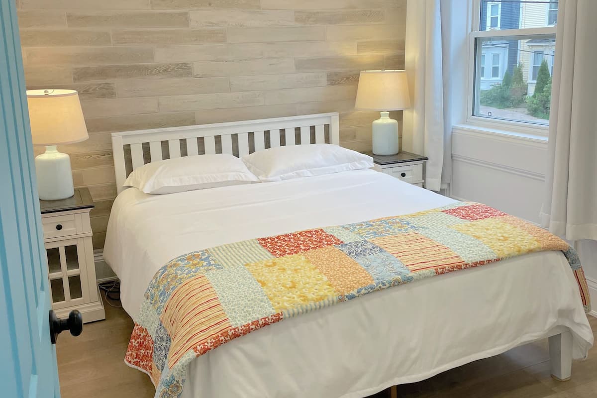 Un dormitorio con cama blanca, cabecera de madera, colcha, dos mesitas de noche con lámparas, una ventana con cortinas y vistas a casas.