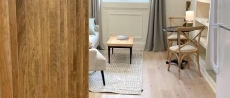 A compact living space with a wooden floor, a dining area with a table and chairs, a sofa, a small rug, and a window with curtains.