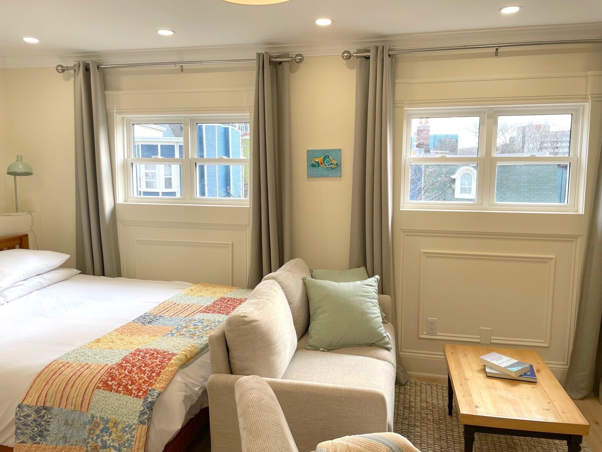 A hotel room with a bed, a sofa, a small table, and two windows with curtains.