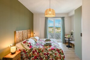 Quarto de hotel com cama, mesa de cabeceira com luminária, varanda com mesa e cadeiras, e vista para a cidade.