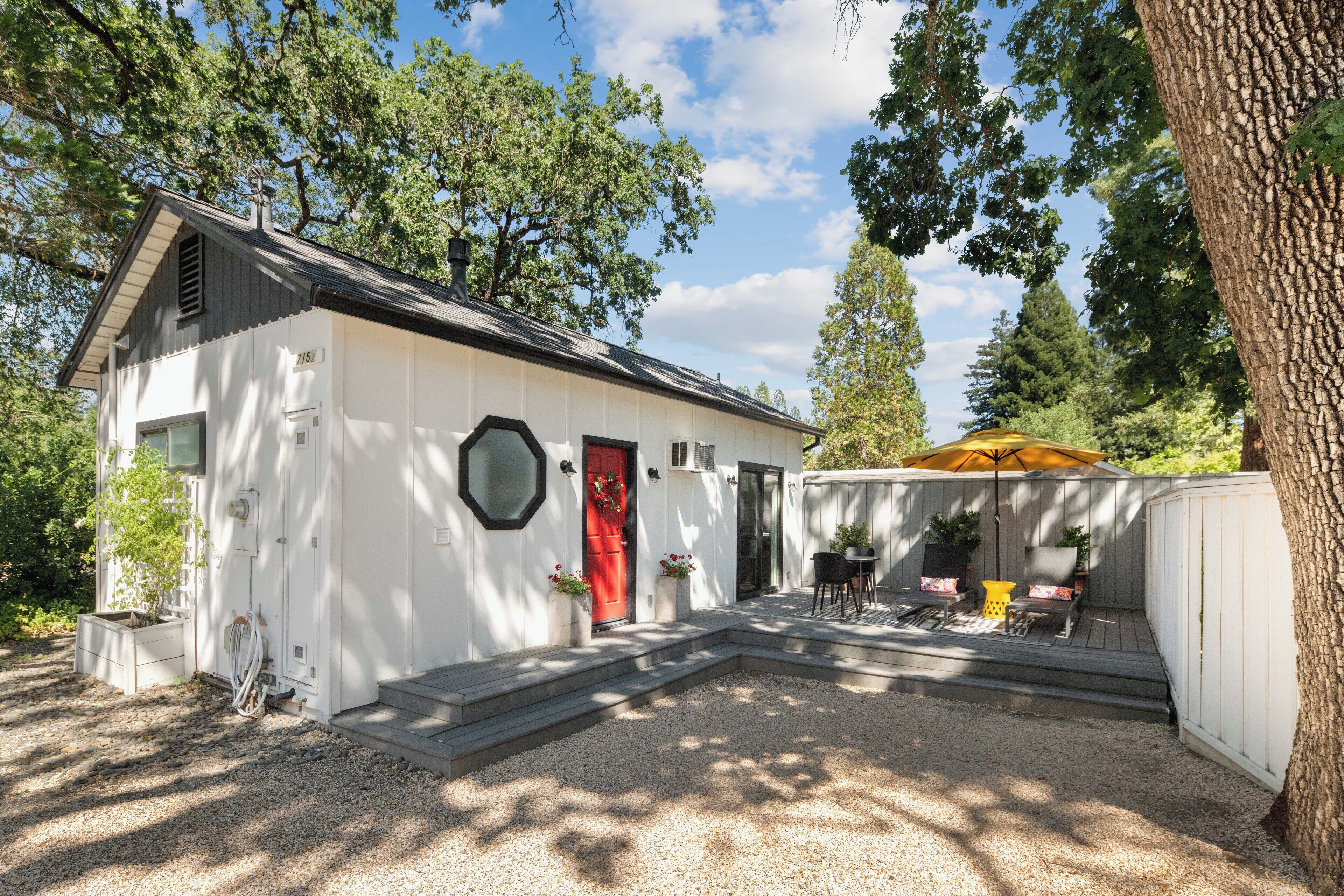 A white building with a red door, a small porch with chairs, and a yellow umbrella.