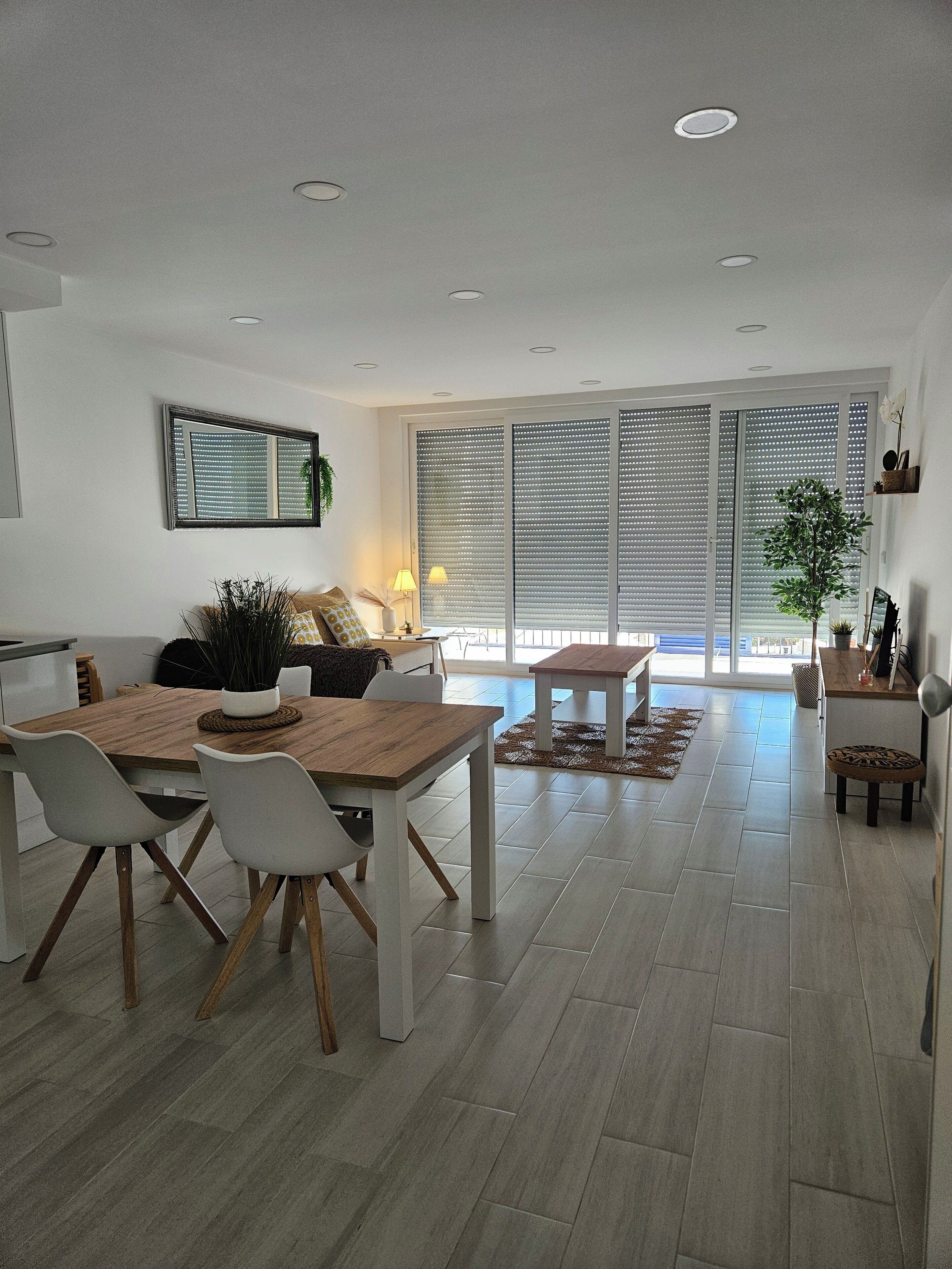 A modern living room with a dining area, featuring a wooden dining table, white chairs, a sofa, and a large window with blinds.