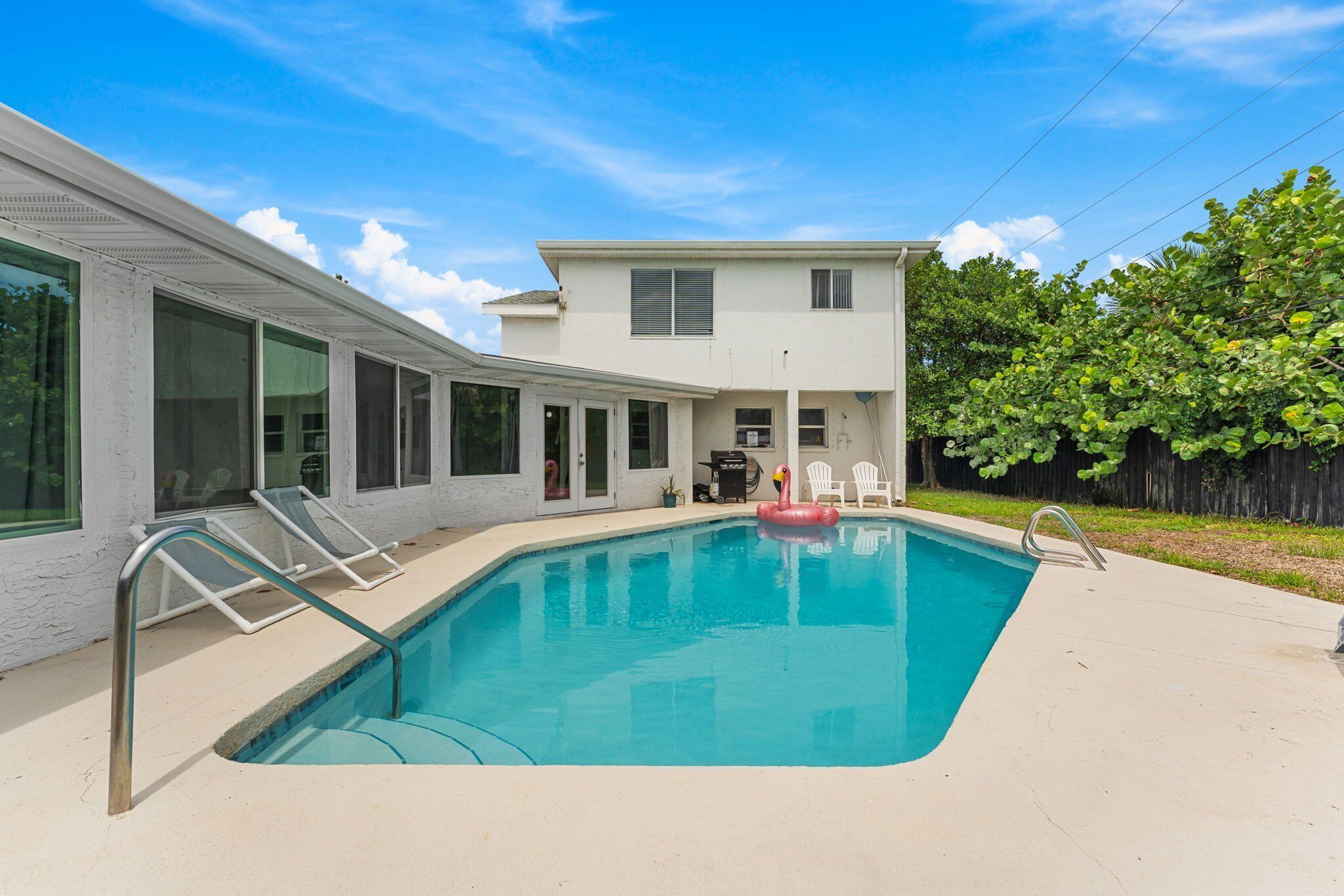 A swimming pool with a clear blue water, surrounded by a concrete deck. There are lounge chairs and a small inflatable flamingo decoration.