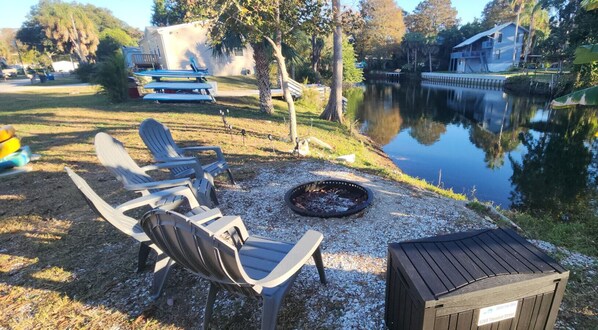 A lakeside outdoor seating area with Adirondack chairs, a fire pit, and a wooden deck. - Shelter from the Storm | Weeki Wachee Riverfront Escape (Weeki Wachee)