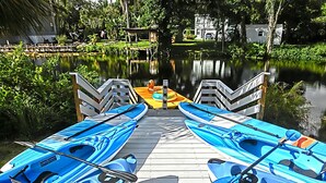 A wooden dock with kayaks lined up on either side, leading into a calm body of water surrounded by greenery. - Shelter from the Storm | Weeki Wachee Riverfront Escape (Weeki Wachee)