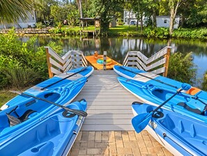 A wooden dock with kayaks lined up, a calm body of water, and greenery in the background.