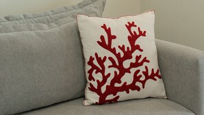Una habitación con un sofá gris, un cojín rojo estampado con motivos de coral, un cuadro colgado en la pared y una ventana.