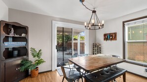 Un coin repas avec une table en bois, des chaises noires et un banc. On y trouve une plante en pot, un meuble en bois avec une enseigne et un porte-bouteilles à vin fixé au mur.