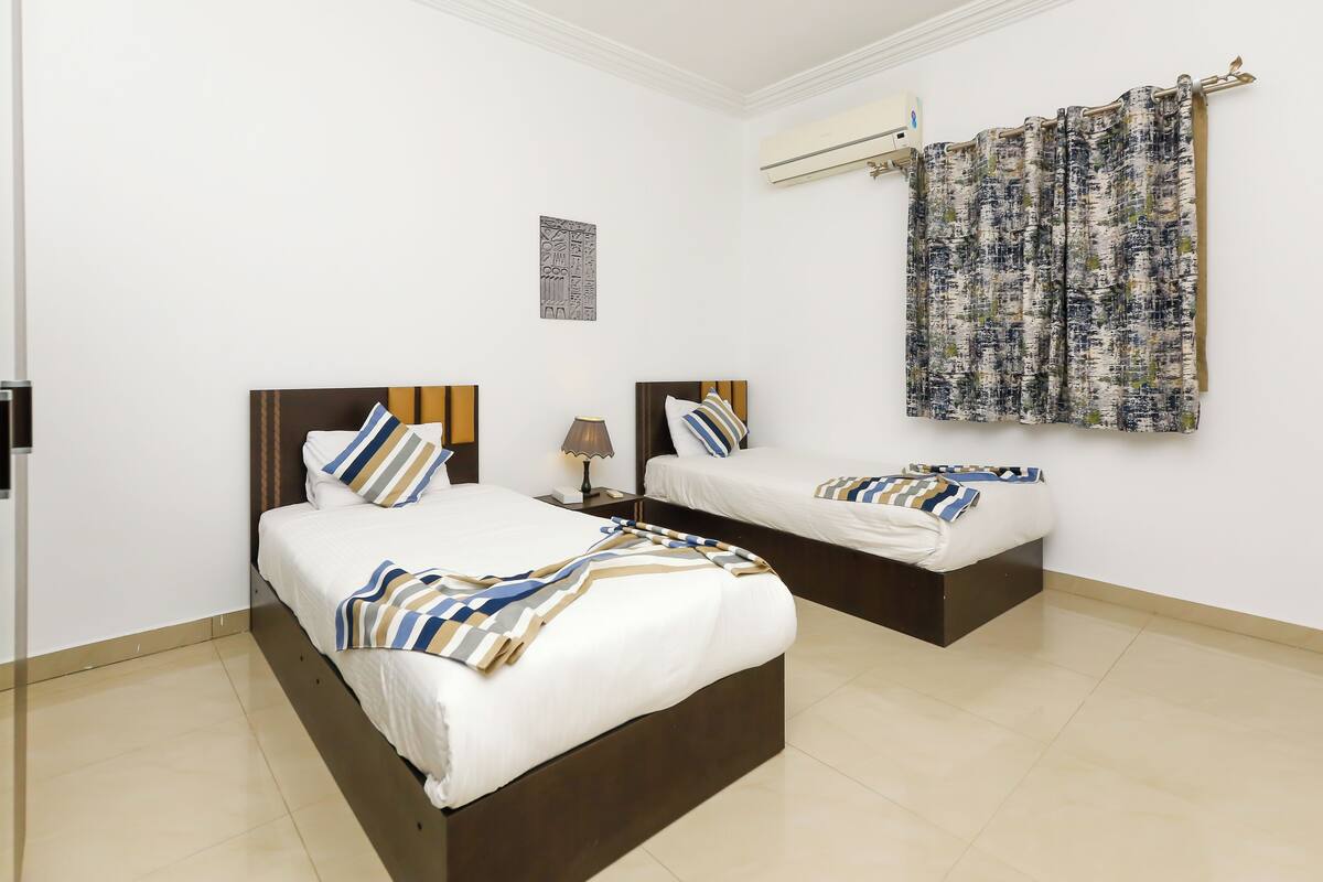 A hotel room with two beds, a nightstand, a lamp, a wall-mounted air conditioner, and a patterned curtain.