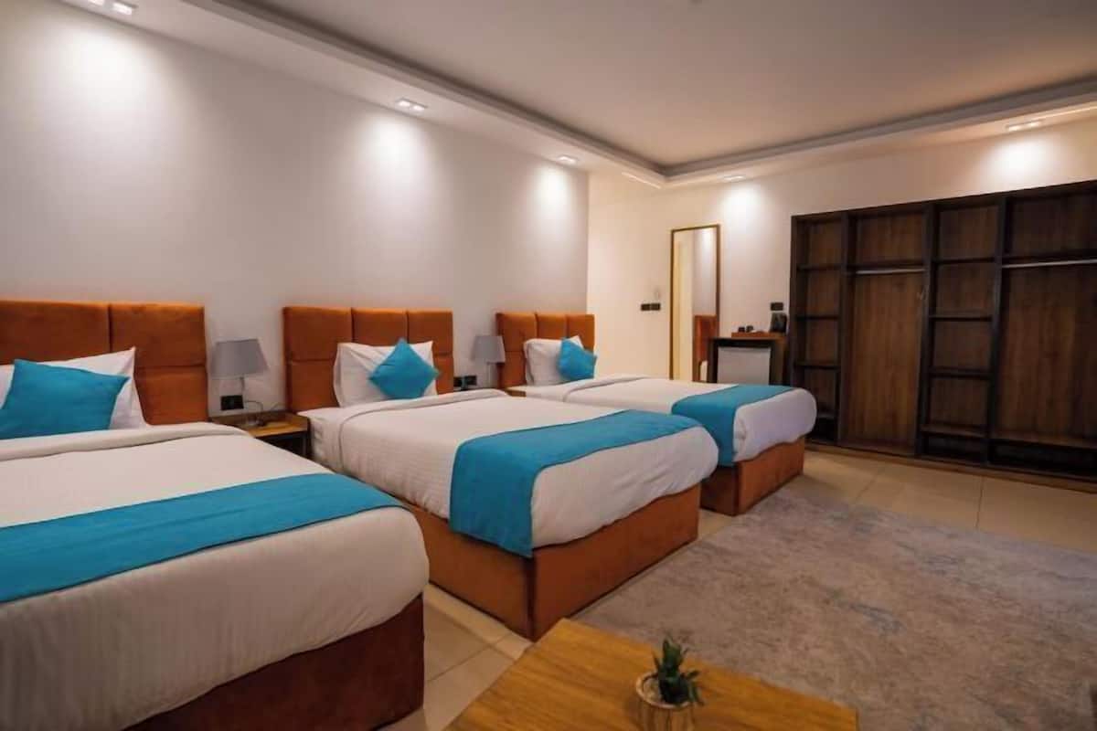 A hotel room with three beds, each with a blue accent pillow, a wooden headboard, and a nightstand with a lamp.