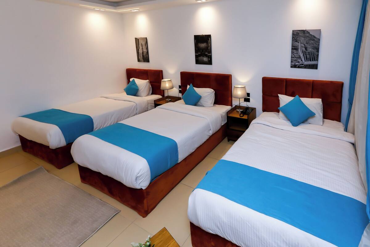 A hotel room with three beds, each with a red headboard, white bedding, and blue accents. There are wall-mounted lamps and artwork above the beds.