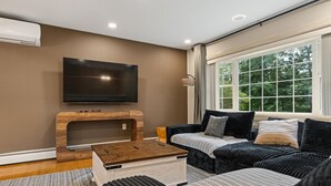 Ein modernes Wohnzimmer mit einem an der Wand befestigten Flachbildfernseher, einem Couchtisch aus Holz, einer gestreiften Couch und einem großen Fenster mit Vorhängen.