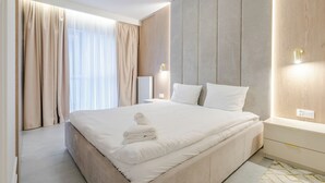 Une chambre d’hôtel moderne avec un grand lit, des rideaux beiges, un mur recouvert de panneaux en bois et une table de chevet avec une lampe.