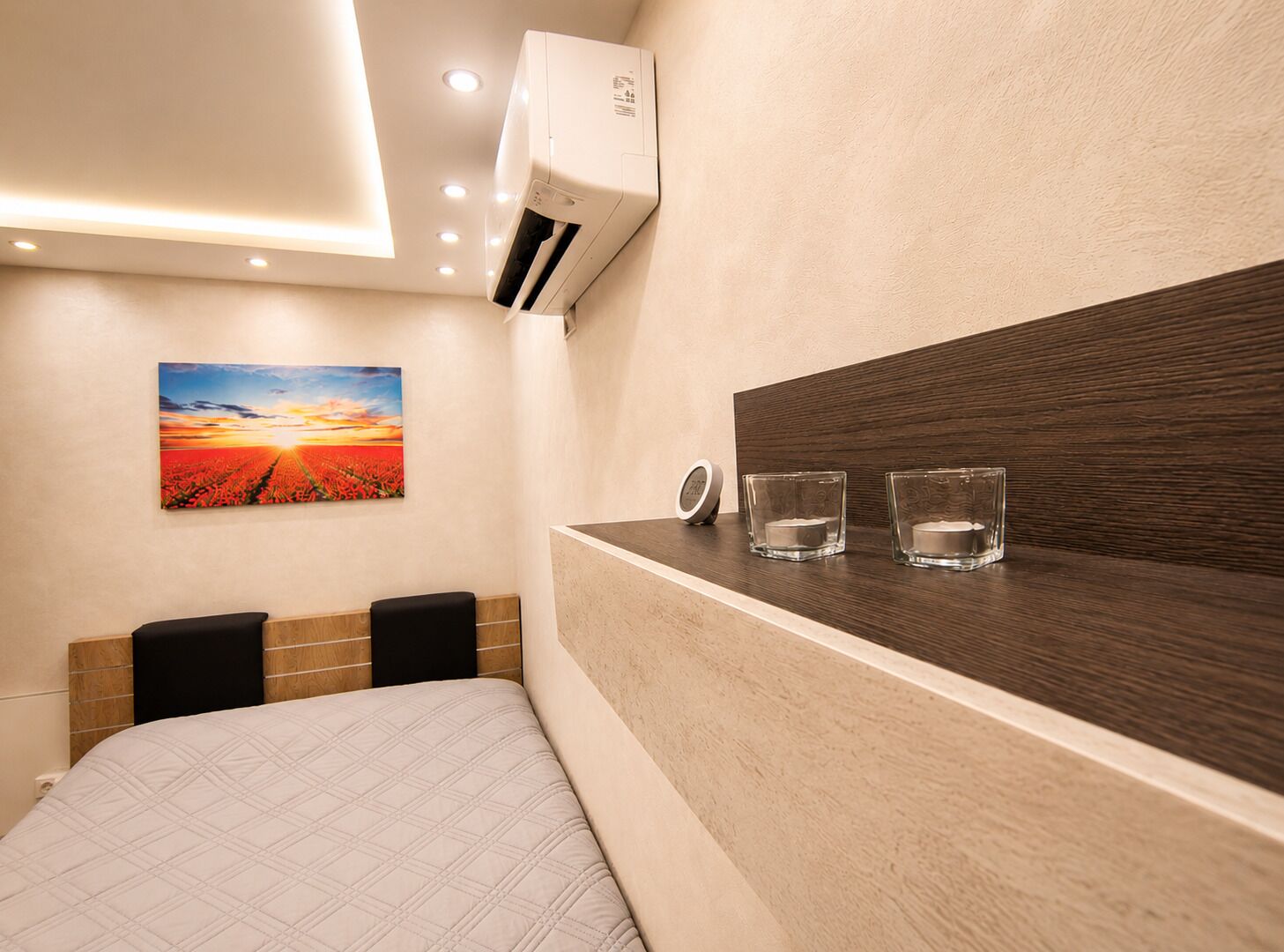 Ein Hotelzimmer mit einem Bett, einer Wand-Klimaanlage, einem Nachttisch mit zwei Gläsern und einem Sonnenuntergangsbild an der Wand.