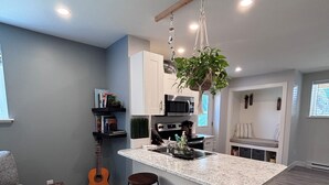 A modern kitchen with a central island, bar stools, a hanging plant, and a guitar. - Private Sauna Retreat • EV Charger • Quiet Creekside Stay (Duncan)