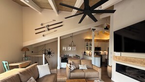 A spacious living area with a high ceiling, a large TV, and a fireplace. There is a kitchen in the background with modern appliances.
