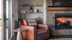 A cozy living room with a leather armchair, a small round table, and a fireplace. Shelves with decorative items and a potted plant are visible. - Luxe | 3 King Suites | Hot Tubs | View | Firepit EV (Banner Elk)