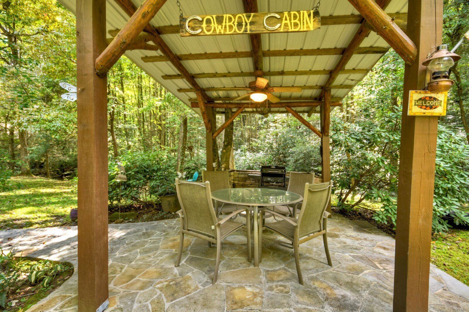 Un área de comedor al aire libre techada con una parrilla de barbacoa, piso de piedra y una estructura de pérgola de madera.