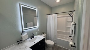 Une salle de bain avec un lavabo blanc, des toilettes, une baignoire avec un rideau de douche et un miroir doté d’un éclairage intégré.