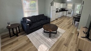 Un salon avec un canapé foncé, une table basse et une petite table d’appoint. On y trouve également un coin cuisine avec des meubles blancs et une table à manger.