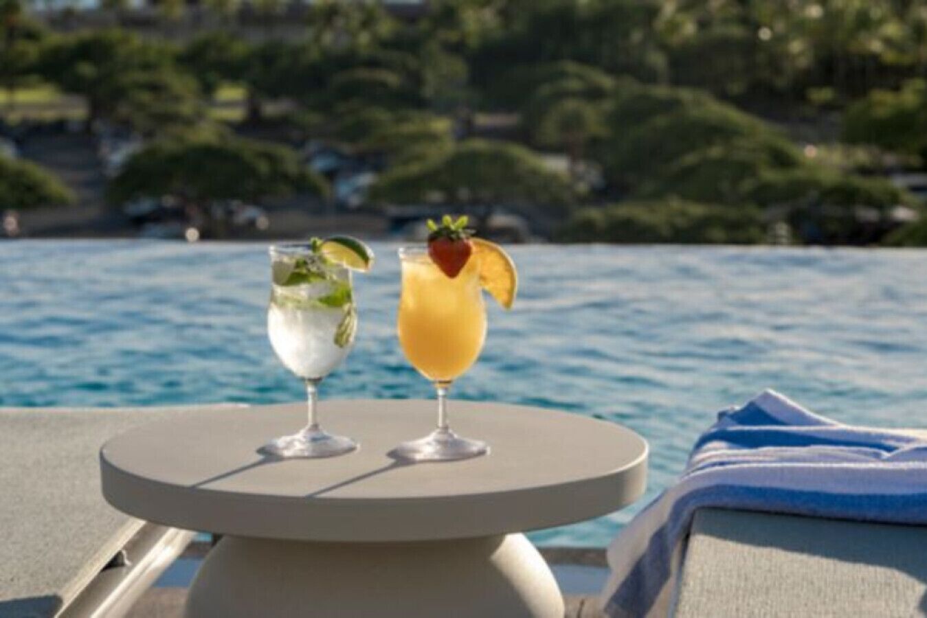 Two drinks on a table by a poolside with a view of the water and greenery in the background.