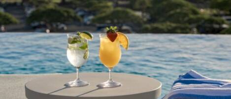 Two drinks on a table by a poolside with a view of the water and greenery in the background.